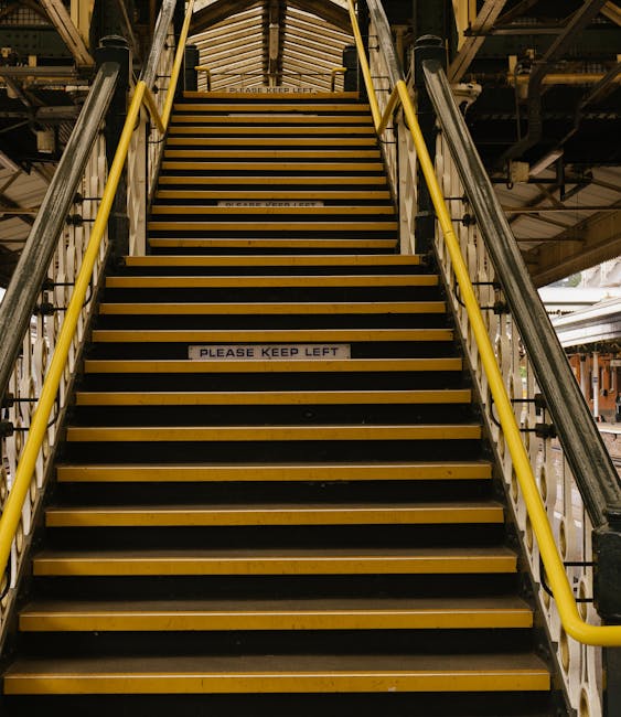 The image shows a set of yellow and black industrial stairs with protective yellow handrails on both sides, leading up to an elevated platform within a warehouse or storage facility. The staircase fea